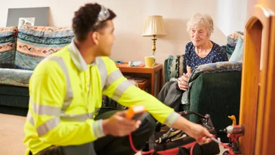Image shows a repairs operative fixing something and an older woman sitting in an armchair
