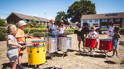 Image shows adults and children playing drums outside