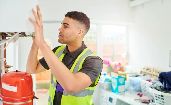 A heating engineer wearing a high-visibility vest inspects a boiler in a laundry room